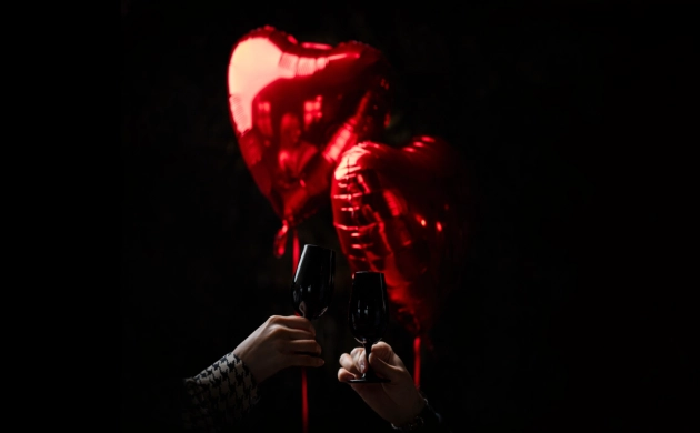 Deux verres de vin trinquant devant des ballons rouges en forme de cœur sur fond sombre, ambiance romantique.