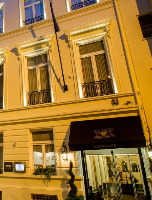 Façade élégante de l'hôtel Stanhope à Bruxelles, éclairée en soirée, avec ses fenêtres encadrées de rideaux et son auvent noir portant le nom de l'établissement.