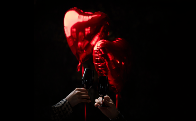 Deux verres de vin trinquant devant des ballons rouges en forme de cœur sur fond sombre, ambiance romantique.